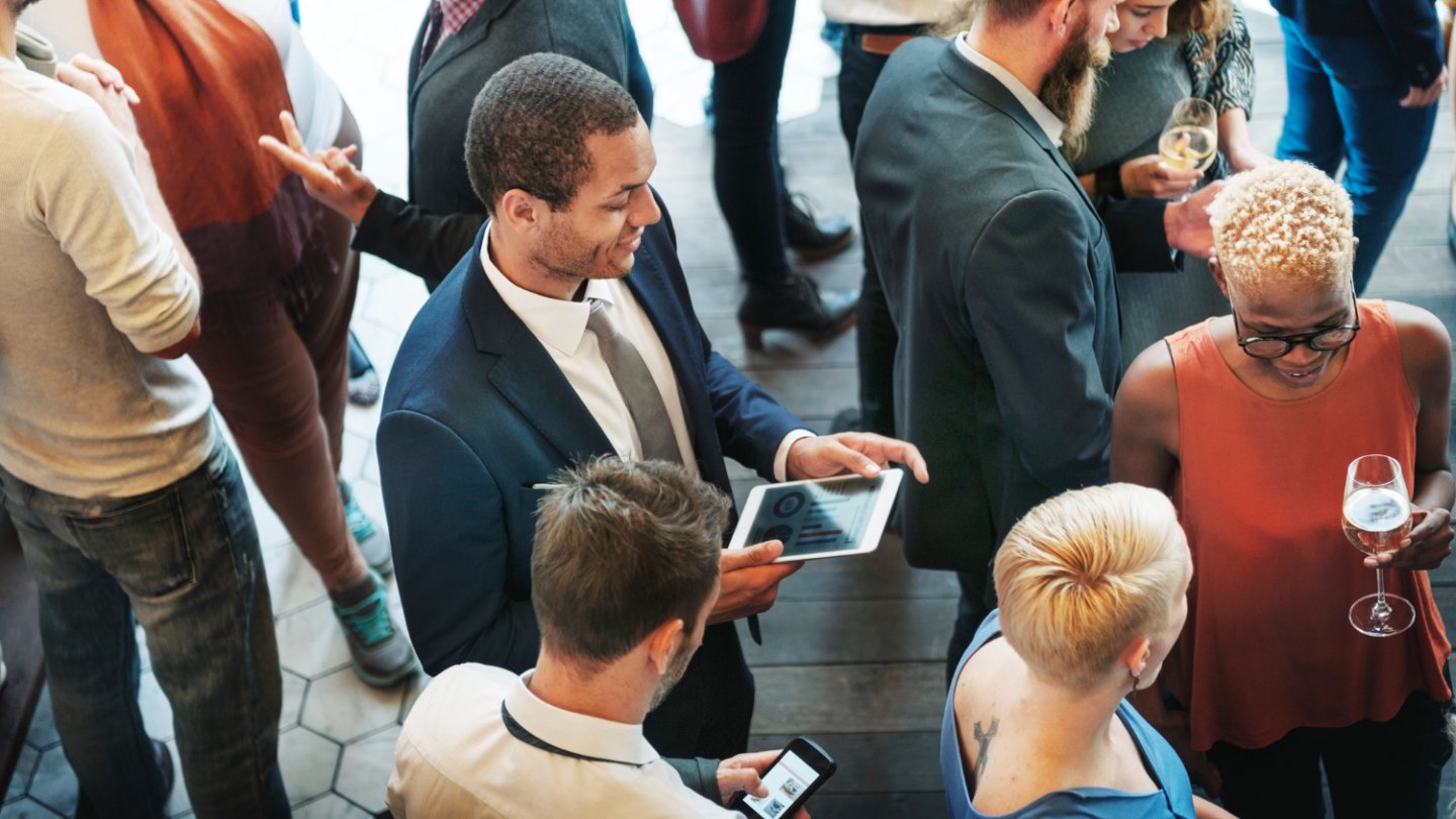 enschen in Business-Kleidung unterhalten sich bei einem Networking-Event in entspannter AtmosphÀre. Einige halten Tablets oder Smartphones, andere ein Glas Wein. Die Szene vermittelt einen professionellen, aber lockeren Austausch unter Veranstaltungsteilnehmenden.