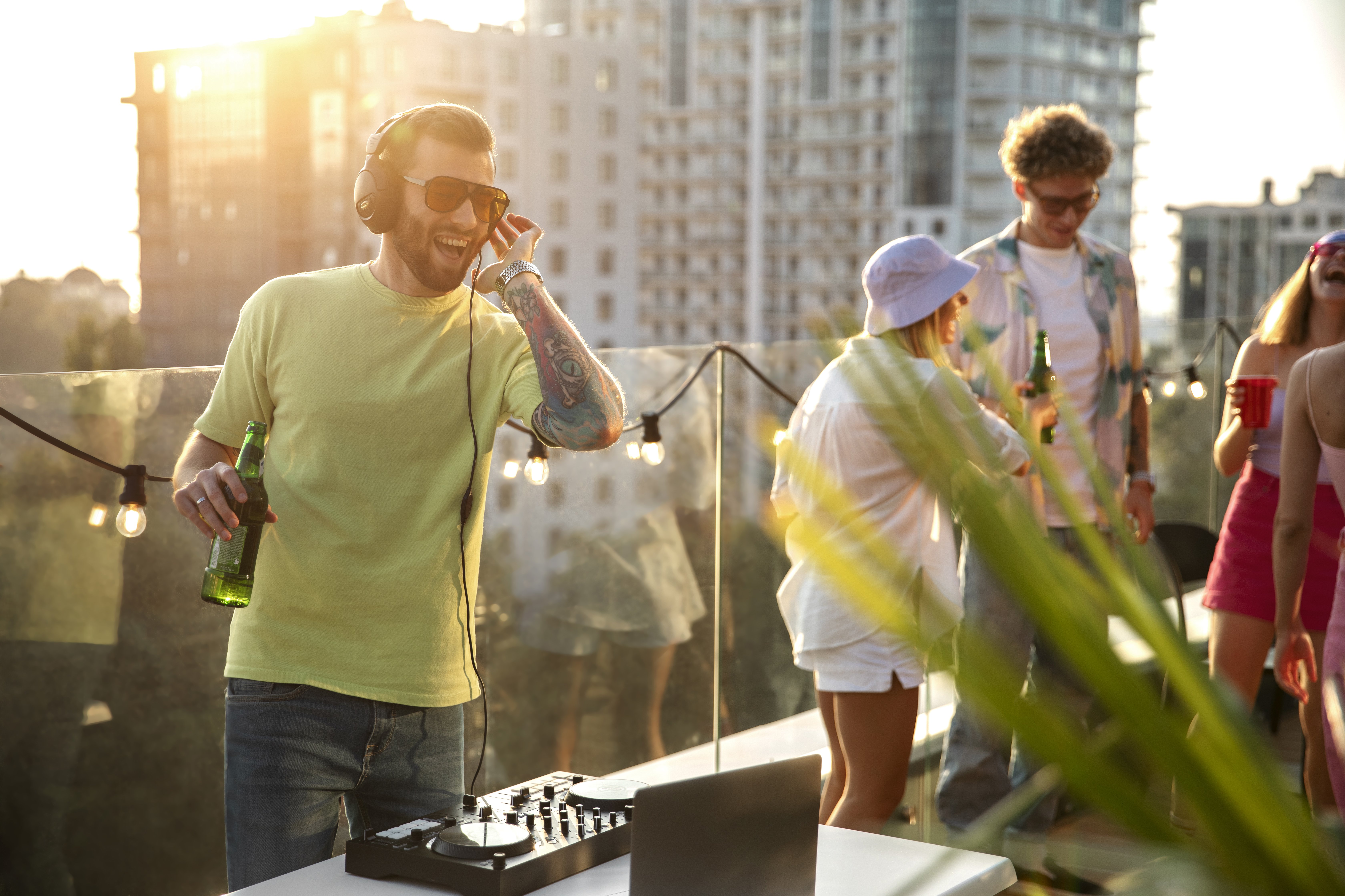 Ein DJ mit Kopfhörern legt bei einer Rooftop-Party im Sonnenuntergang auf, während Freunde um ihn herum tanzen und sich unterhalten.