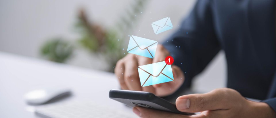 Person hÀlt ein Smartphone in der Hand, auf dem eine neue E-Mail-Benachrichtigung angezeigt wird; im Hintergrund ist eine Computertastatur zu sehen.