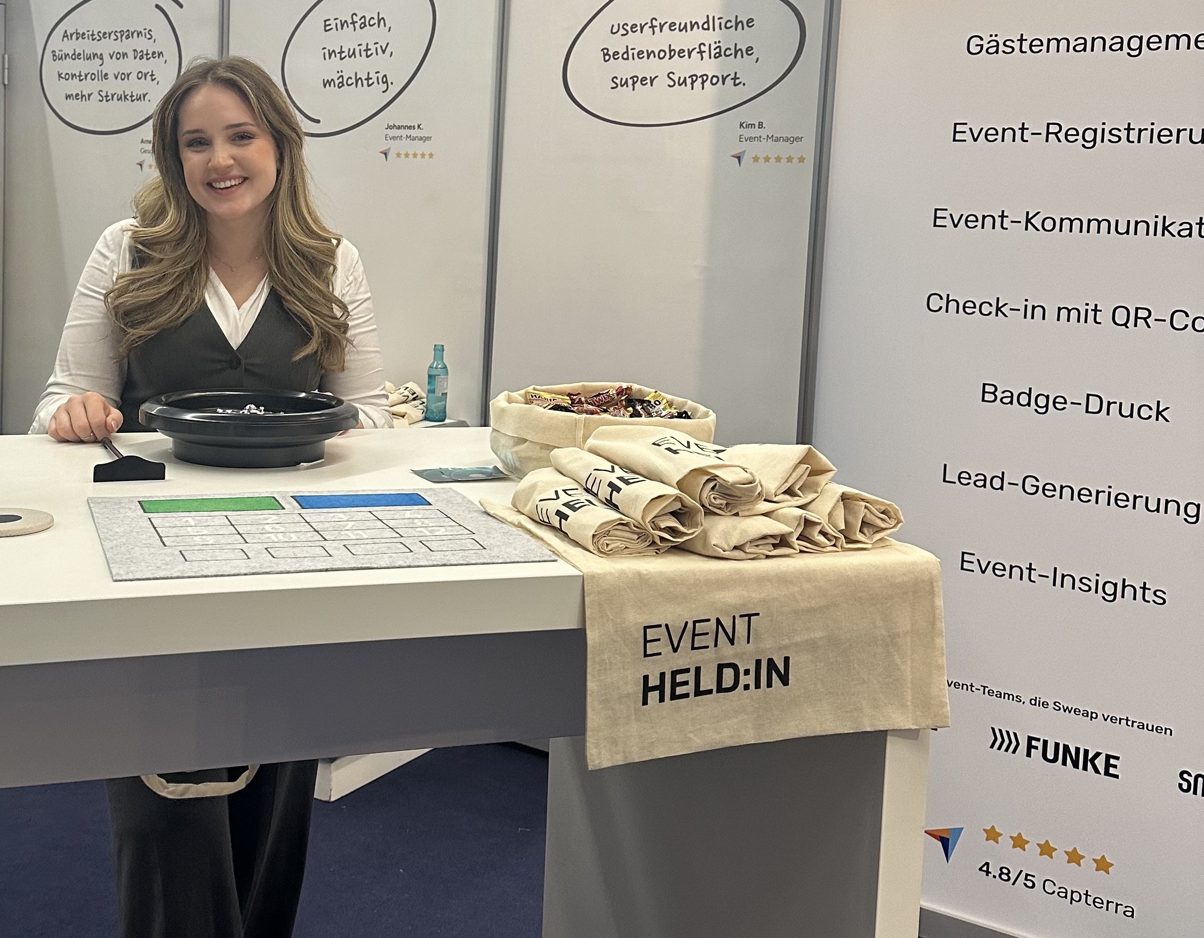 Mia from Sweap presents a recycled cotton tote bag with the words 'EVENT HERO' as a sustainable trade show giveaway at a booth.