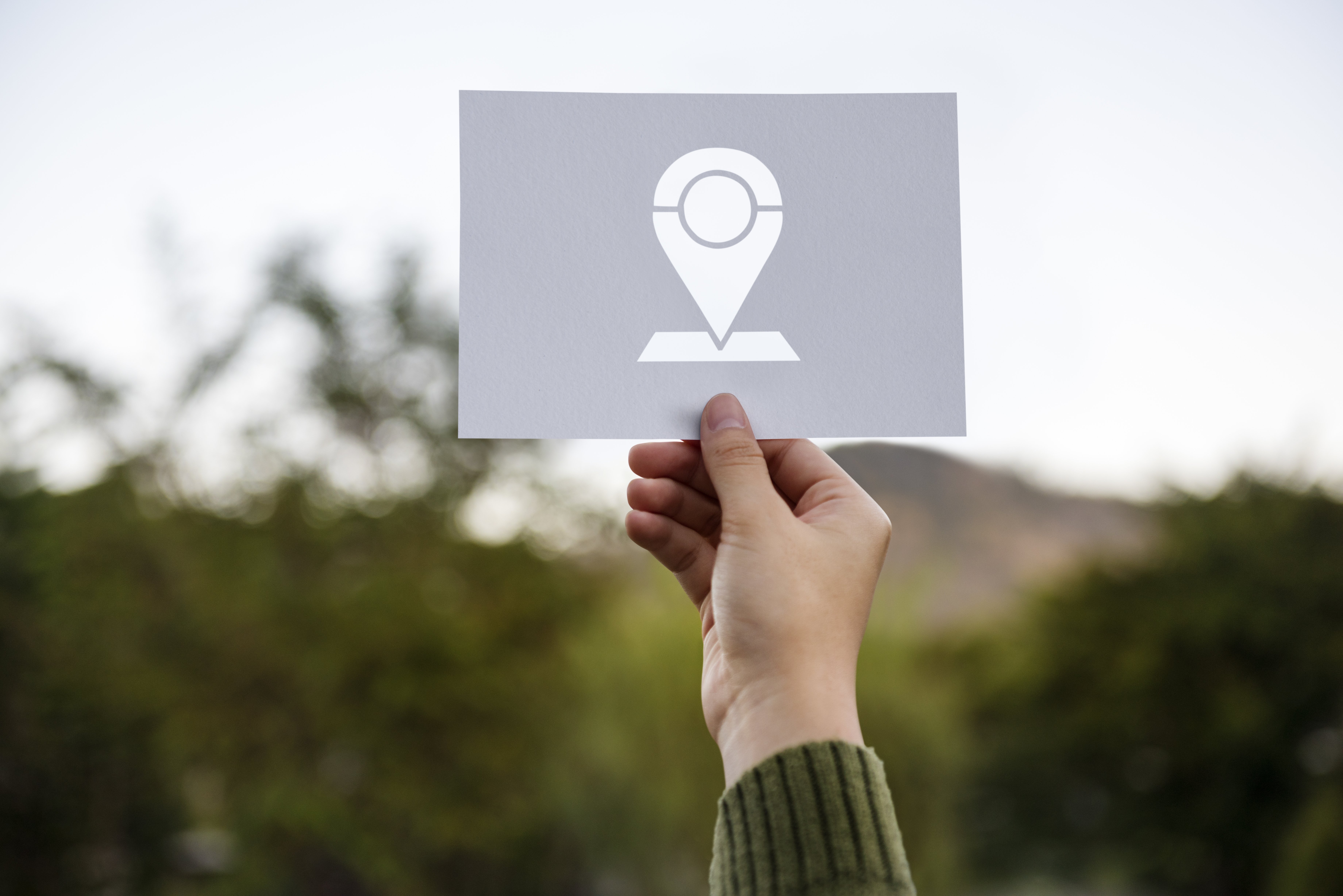 A hand holds up a sign with a location pin icon against a blurry natural landscape.