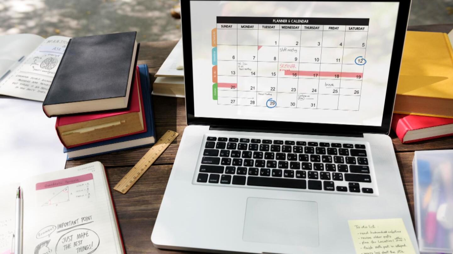 Laptop with an open calendar planner on a wooden desk, surrounded by notebooks, pens, and rulers – planning environment for an event or project.
