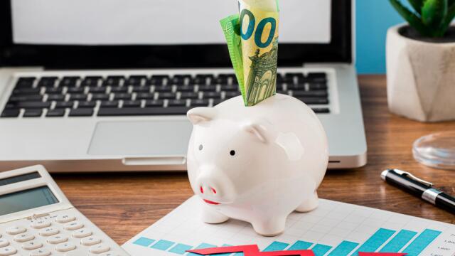 A white piggy bank with 100-euro bills sticking out sits on a desk in front of a laptop. A calculator, a chart with a red arrow, and a plant are nearby—symbolizing financial planning and budget optimization.