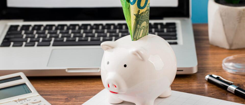 A white piggy bank with 100-euro bills sticking out sits on a desk in front of a laptop. A calculator, a chart with a red arrow, and a plant are nearby—symbolizing financial planning and budget optimization.