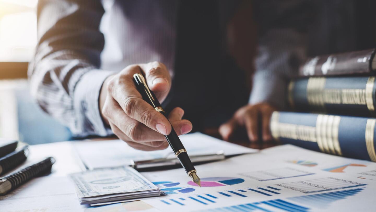 Business person analyzing financial charts and budget documents at a desk, with stacks of cash, graphs, and law books in the background.