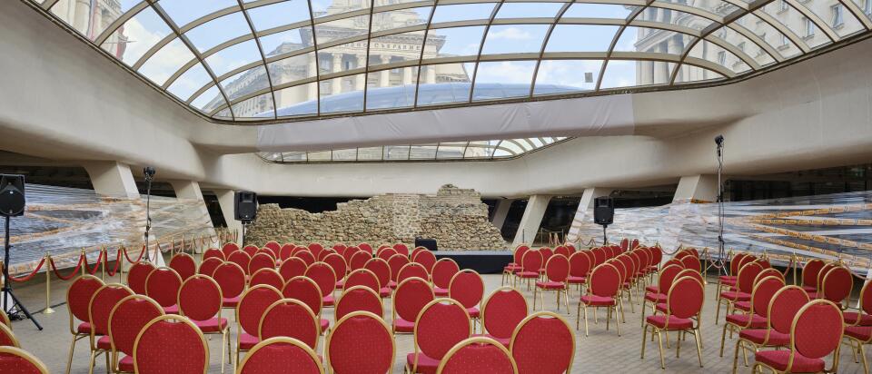 Ein Veranstaltungssaal unter einem gewölbten Glasdach mit Reihen roter Stühle, die auf eine antike Steinruine ausgerichtet sind; durch das Dach sind historische Gebäude sichtbar.