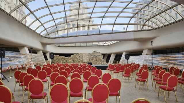 Ein Veranstaltungssaal unter einem gewölbten Glasdach mit Reihen roter Stühle, die auf eine antike Steinruine ausgerichtet sind; durch das Dach sind historische Gebäude sichtbar.