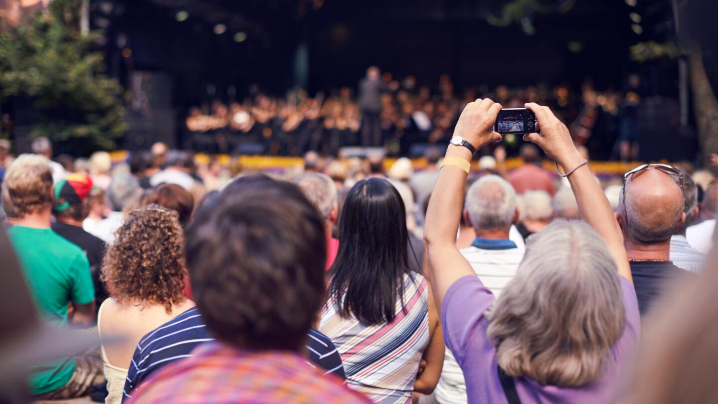 Crowd at an outdoor event with one person holding up a smartphone to take a photo.
