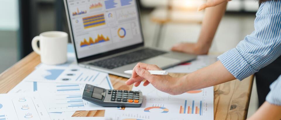 Nahaufnahme von Händen, die einen Taschenrechner auf einem Schreibtisch voller Finanzdiagramme und einem Laptop mit Datenvisualisierungen nutzen.