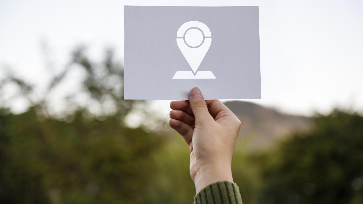 Eine Hand hält ein Schild mit einem Standort-Symbol vor eine unscharfe Naturlandschaft.