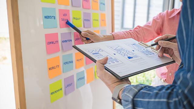 Marketing-Team stimmt Aufgaben an einem Whiteboard mit Post-its und Entwürfen für ein strukturiertes Projekt-Management ab.