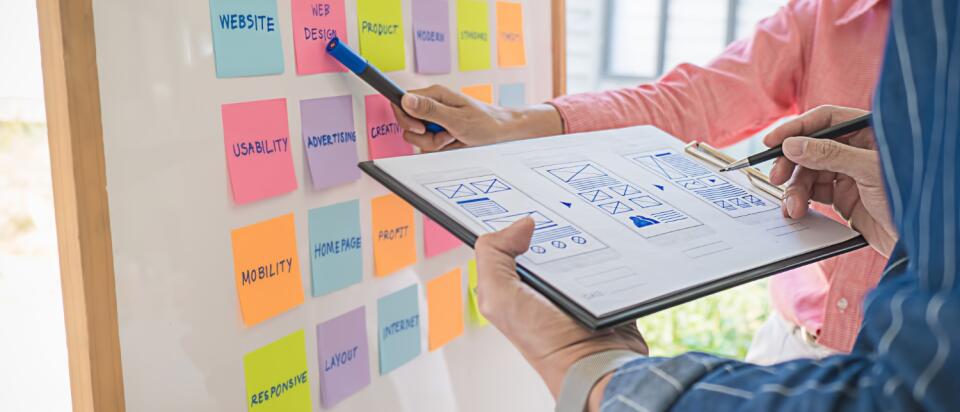 Marketing-Team stimmt Aufgaben an einem Whiteboard mit Post-its und Entwürfen für ein strukturiertes Projekt-Management ab.