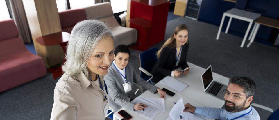 Overhead view of a woman with a marker in front of a whiteboard, while a team wearing conference badges works together at a table with a laptop and documents in the background