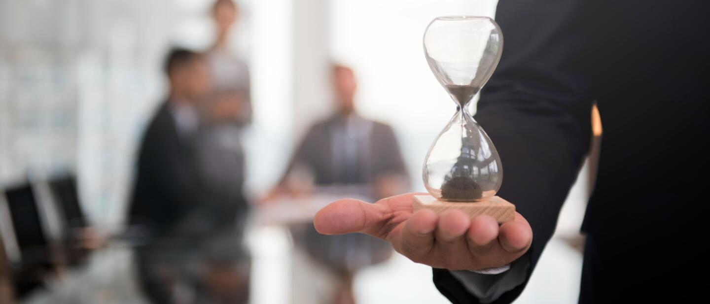 Geschäftsmann hält eine Sanduhr in der Hand, während im Hintergrund verschwommen ein Team bei einer Besprechung zu sehen ist – Symbol für Zeitmanagement bei Events.