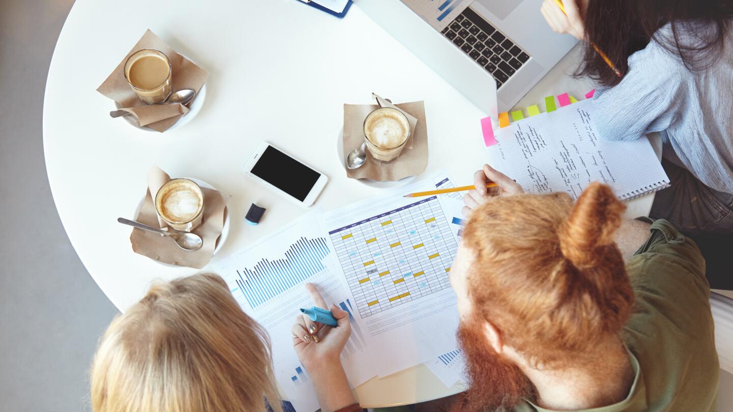 Blick von oben auf einen Tisch mit Planungsunterlagen, Diagrammen und Kalender, umgeben von Kaffeetassen und Notizen. Drei Personen arbeiten gemeinsam an der Auswertung
