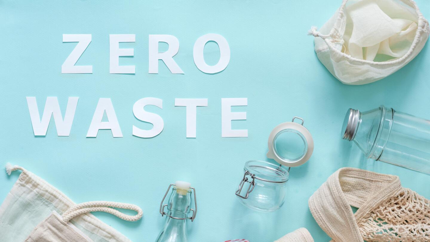 Der Schriftzug “ZERO WASTE” liegt auf einem hellblauen Hintergrund, umgeben von wiederverwendbaren Glasflaschen, Gläsern und Stoffbeuteln.
