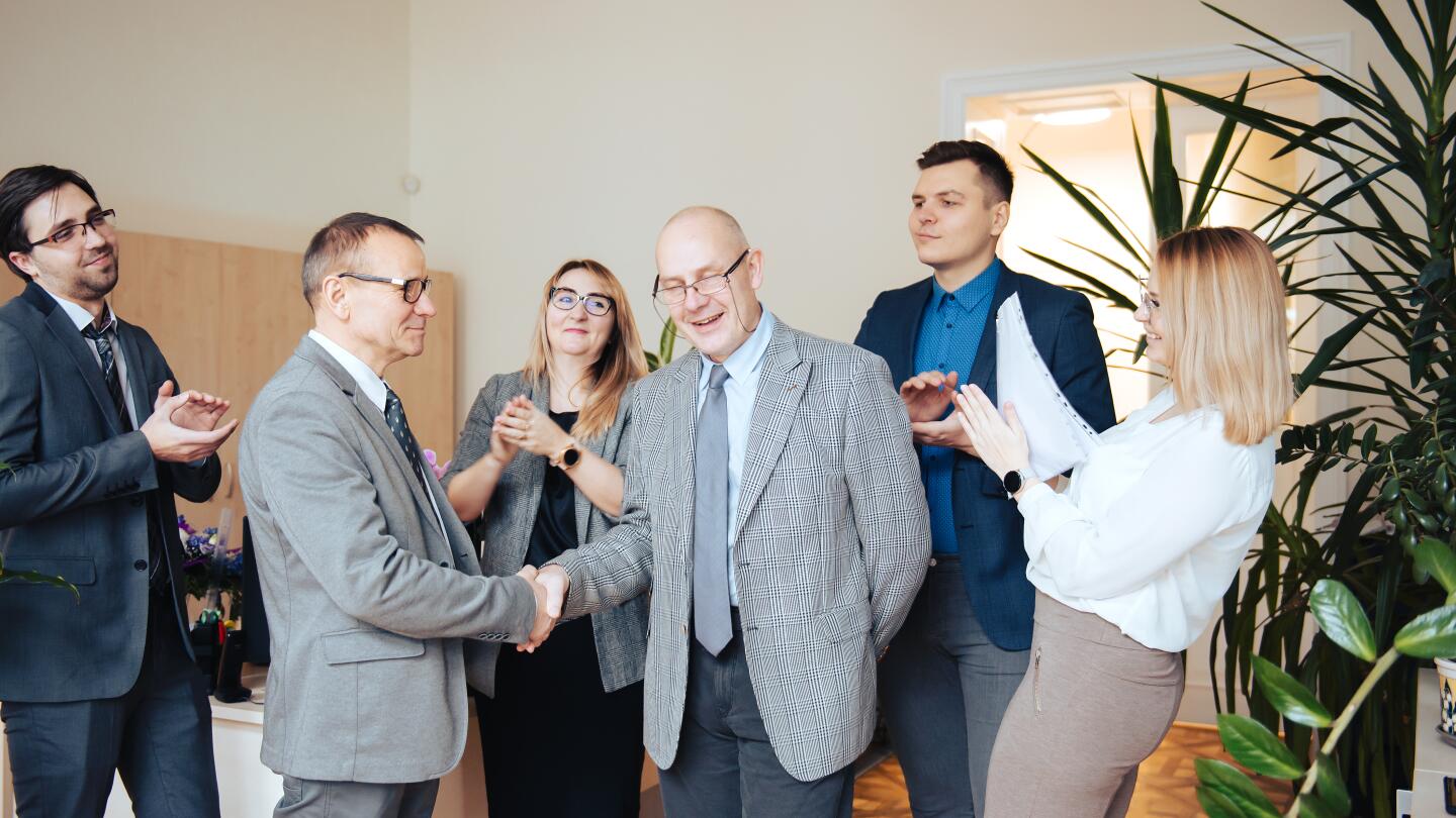 Zwei Geschäftsmänner schütteln sich lächelnd die Hand, während umstehende Kollegen applaudieren – Symbolbild für einen erfolgreichen Geschäftsabschluss