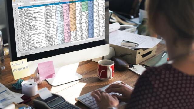 Eine Person arbeitet an einem Schreibtisch an einer “Marketing Budget”-Tabelle auf einem großen Computermonitor, umgeben von Büromaterial.
