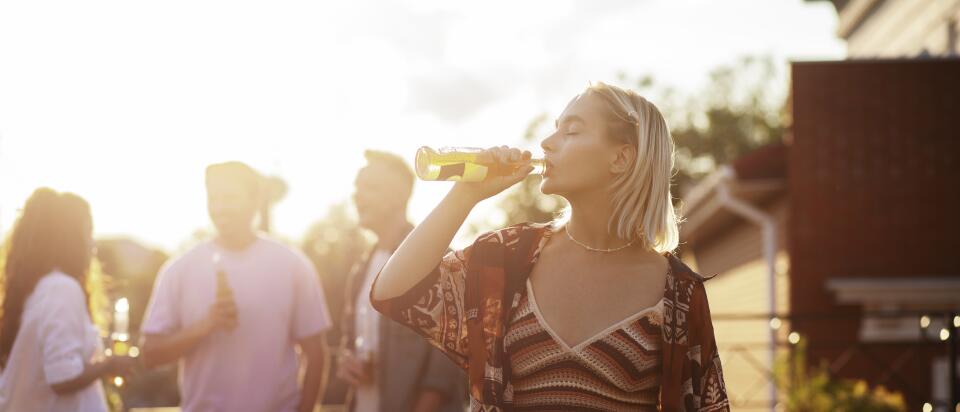 Junge Frau trinkt aus einer Flasche bei einer lockeren Dachterrassen-Party im Sonnenuntergang, im Hintergrund weitere Gäste mit Getränken