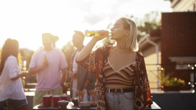 Junge Frau trinkt aus einer Flasche bei einer lockeren Dachterrassen-Party im Sonnenuntergang, im Hintergrund weitere Gäste mit Getränken